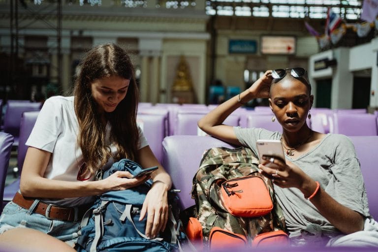 Deux voyageuses assises avec sacs à dos consultant leurs smartphones en salle d'attente