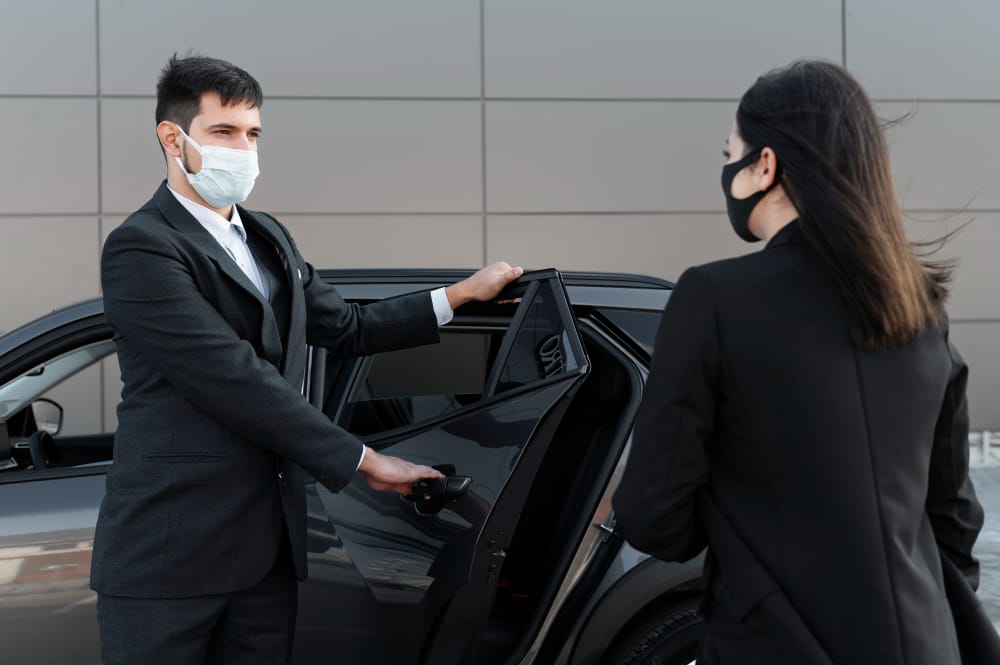 Chauffeur masqué ouvre la porte d’une berline à une passagère